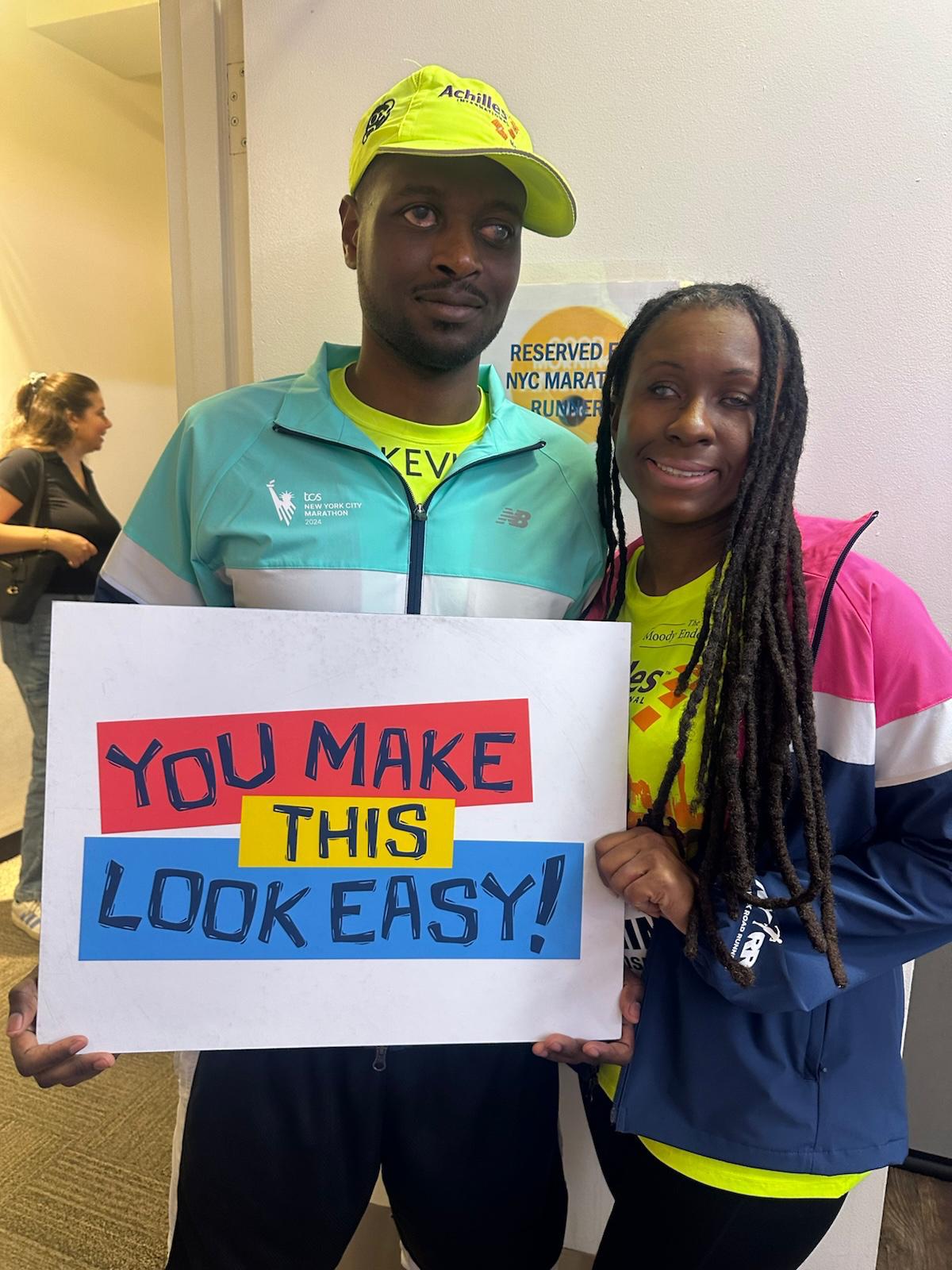 Kevin and Jasmine at the 2024 New York City Marathon, wearing their Team Inspire jackets and holding a sign that reads: You make this look easy!
